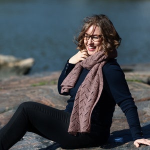 May include: A woman wearing a navy blue sweater and black jeans sits on a rocky surface. She is wearing a brown knitted scarf and glasses. She is smiling and looking to the side.