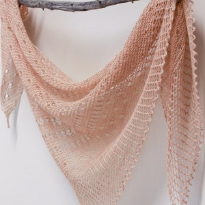 May include: A peach-coloured triangular shawl, with an open, lacy knit design and a scalloped edge, is draped over a weathered branch against a white backdrop. The shawl's texture and colour suggest a handmade item. The branch is a natural, greyish-brown colour.