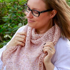 May include: A woman wearing a white shirt and glasses holds a pink crocheted shawl with a delicate lace pattern.