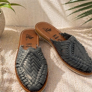 May include: A pair of dark grey woven leather huarache sandals with a tan leather insole. The insole features a printed cactus logo and text that reads MEX 24 USA 7. The sandals have a closed-toe design with intricate braided leather straps.