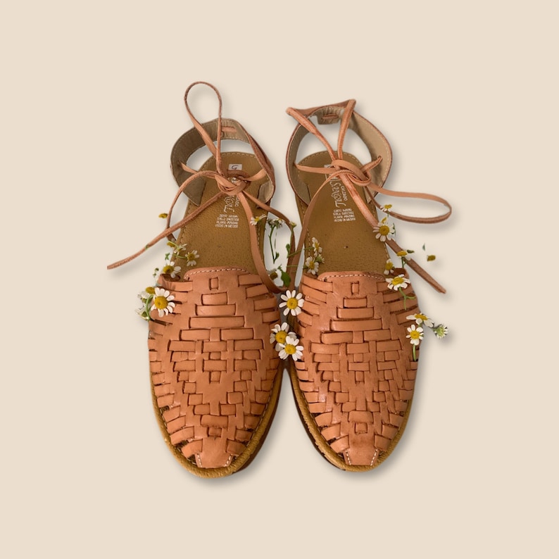 Puede incluir: Un par de sandalias huarache de color tostado con empeine de cuero trenzado, dise&ntilde;o de punta cerrada y cordones largos para el tobillo. Peque&ntilde;as flores de margarita blancas con centro amarillo est&aacute;n insertadas en las tiras de cuero trenzado.