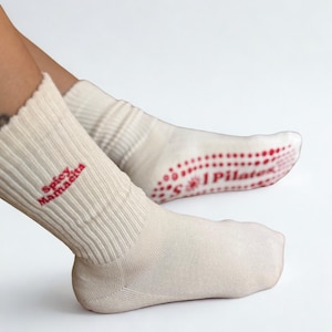 May include: Cream-coloured Pilates socks with red non-slip grips on the soles. The left sock has the text "Spicy Mamaeita" in red, while the right sock features "Pilates" in red. The socks are ribbed at the ankle.