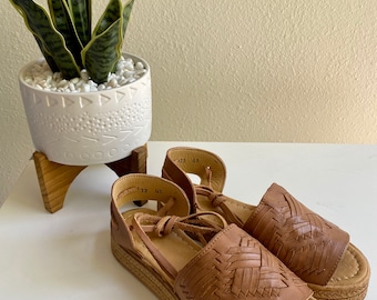 May include: A pair of tan leather huarache sandals featuring a woven upper design, an open toe, and a wrap-around ankle strap. The sandals have a thick, textured platform sole with a braided jute-style trim around the edge of the base.