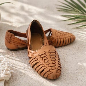 May include: A pair of tan leather huarache sandals featuring a woven lattice design across the upper, a closed rounded toe, and an open heel construction with a flat sole. The leather has a matte finish and a natural light brown color.