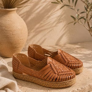 Op de afbeelding: Een paar bruine leren huarache-schoenen met een gevlochten bovenwerk, een gesloten ronde neus en een dikke plateauzool omwikkeld met natuurlijke jutvezel. De binnenkant van de schoen is gevoerd met bruin leer en bevat zichtbare maatmarkeringen.