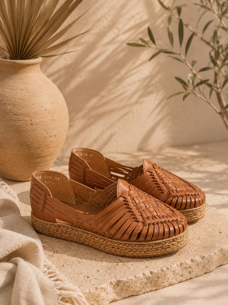 Op de afbeelding: Een paar bruine leren huarache-schoenen met een gevlochten bovenwerk, een gesloten ronde neus en een dikke plateauzool omwikkeld met natuurlijke jutvezel. De binnenkant van de schoen is gevoerd met bruin leer en bevat zichtbare maatmarkeringen.
