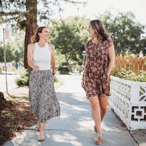 Op de afbeelding: Een wit mouwloos tanktop met racerback gecombineerd met een zwart-witte midi-rok met luipaardprint, en een bruine jurk met korte mouwen en een wit bloemenpatroon, beide gedragen met platte sandalen tijdens het lopen op een stoep.