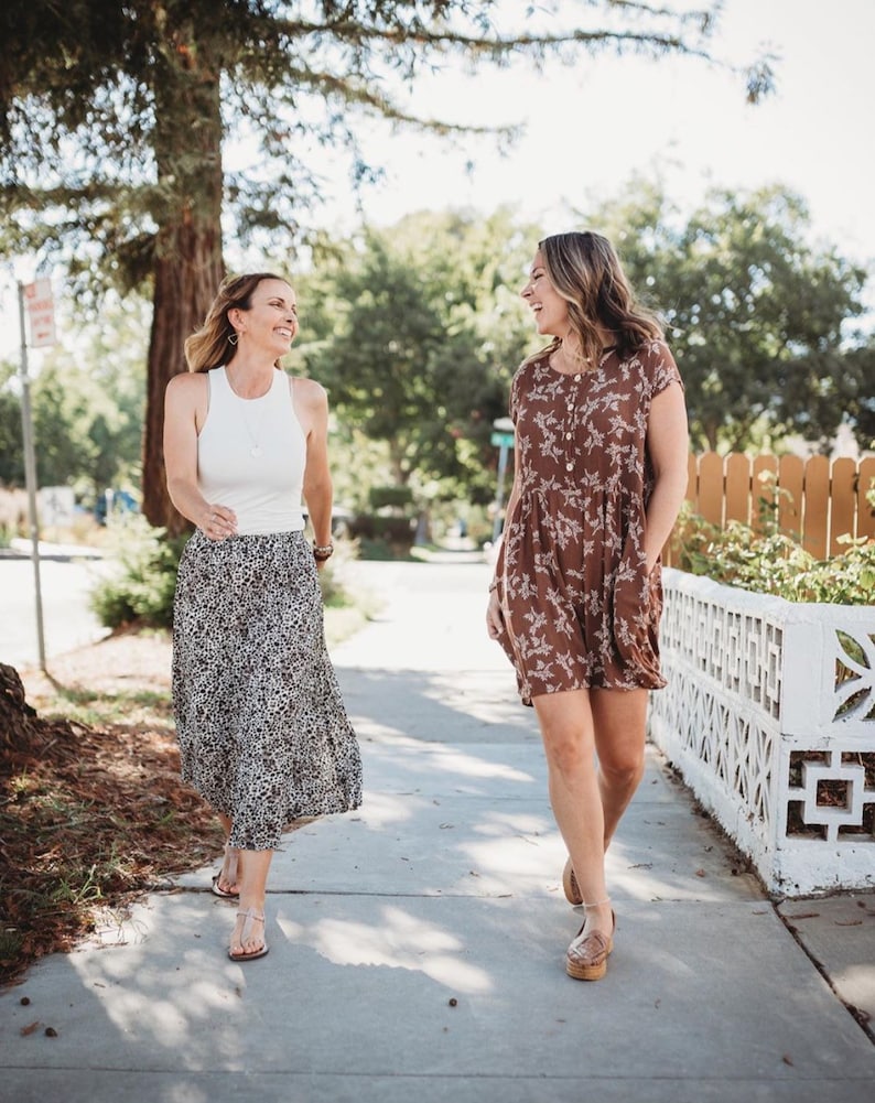 Op de afbeelding: Een wit mouwloos tanktop met racerback gecombineerd met een zwart-witte midi-rok met luipaardprint, en een bruine jurk met korte mouwen en een wit bloemenpatroon, beide gedragen met platte sandalen tijdens het lopen op een stoep.