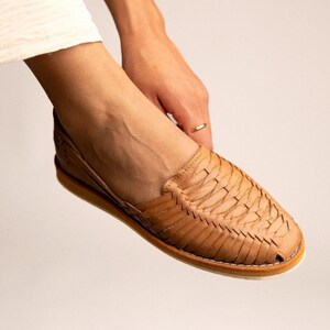 Peut inclure: Une chaussure huarache beige dot&eacute;e d'une tige en cuir tress&eacute; avec un bout ferm&eacute; et une semelle plate. La chaussure pr&eacute;sente un style &agrave; enfiler avec des bandes de cuir entrelac&eacute;es formant un motif textur&eacute; sur le dessus et les c&ocirc;t&eacute;s.
