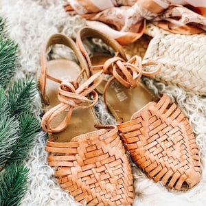 Puede incluir: Un par de sandalias huarache de cuero color canela con un dise&ntilde;o de tejido entrelazado en la parte superior, punta cerrada y larga, y cordones de cuero que se atan alrededor del tobillo. Las sandalias tienen una suela plana y tal&oacute;n estructurado.