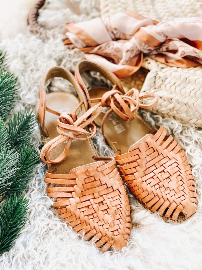 Puede incluir: Un par de sandalias huarache de cuero color canela con un dise&ntilde;o de tejido entrelazado en la parte superior, punta cerrada y larga, y cordones de cuero que se atan alrededor del tobillo. Las sandalias tienen una suela plana y tal&oacute;n estructurado.