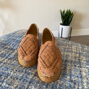 Op de afbeelding: Een paar lichtbruine instapschoenen met een bovenwerk van gevlochten leer en een dikke, getextureerde plateauzool. De schoenen hebben een ronde neus en een open constructie, met een gedetailleerd vlechtpatroon over de voorzijde van de schoen.