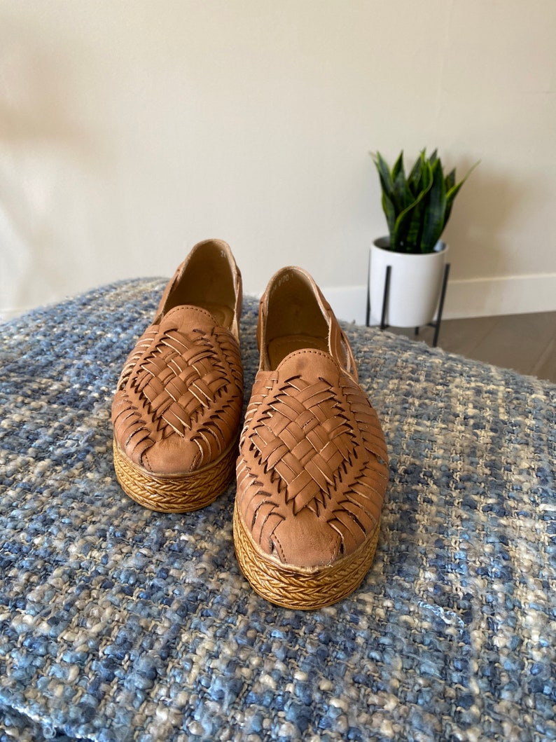 Op de afbeelding: Een paar lichtbruine instapschoenen met een bovenwerk van gevlochten leer en een dikke, getextureerde plateauzool. De schoenen hebben een ronde neus en een open constructie, met een gedetailleerd vlechtpatroon over de voorzijde van de schoen.