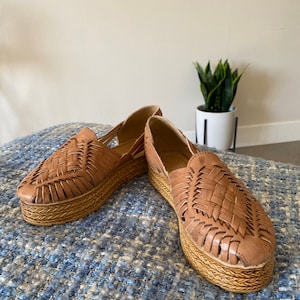 Op de afbeelding: Een paar schoenen in huarache-stijl van beige leer met een gevlochten bovenwerk en een dikke, gevlochten jute plateauzool. De schoenen hebben een gesloten neus met zijdelingse uitsparingen en een instapmodel silhouet.