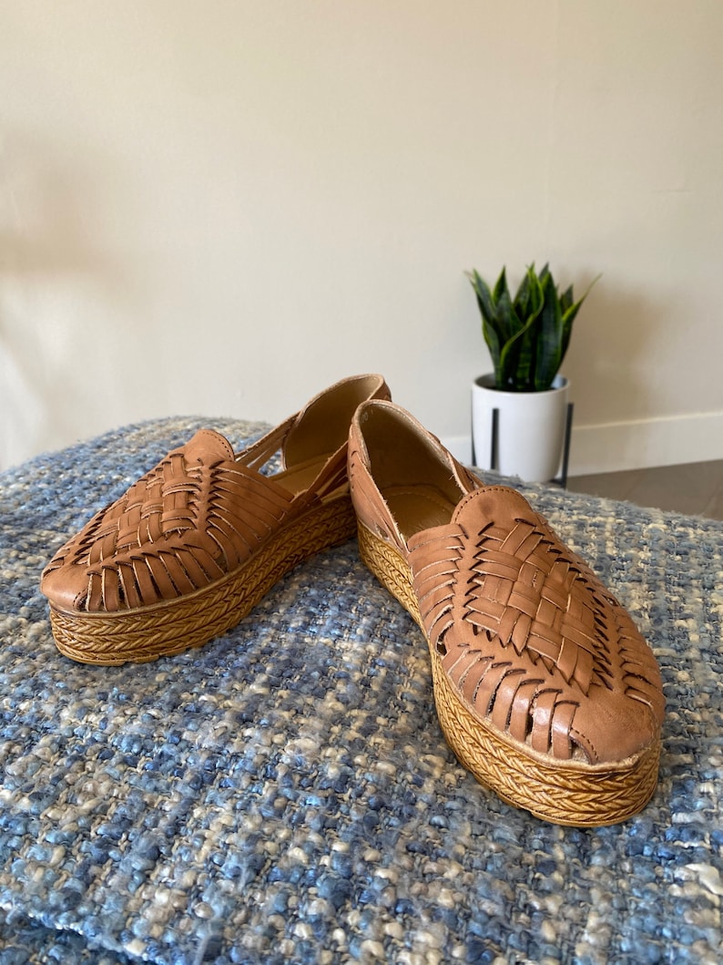 Op de afbeelding: Een paar schoenen in huarache-stijl van beige leer met een gevlochten bovenwerk en een dikke, gevlochten jute plateauzool. De schoenen hebben een gesloten neus met zijdelingse uitsparingen en een instapmodel silhouet.