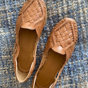 Op de afbeelding: Een paar bruine leren huarache-instappers met een gevlochten bovenwerk, een gesloten neus en een open zijkant. De schoenen hebben een gladde leren binnenzool en een flexibel, laag profiel met gedetailleerde handgeweven leren stroken.
