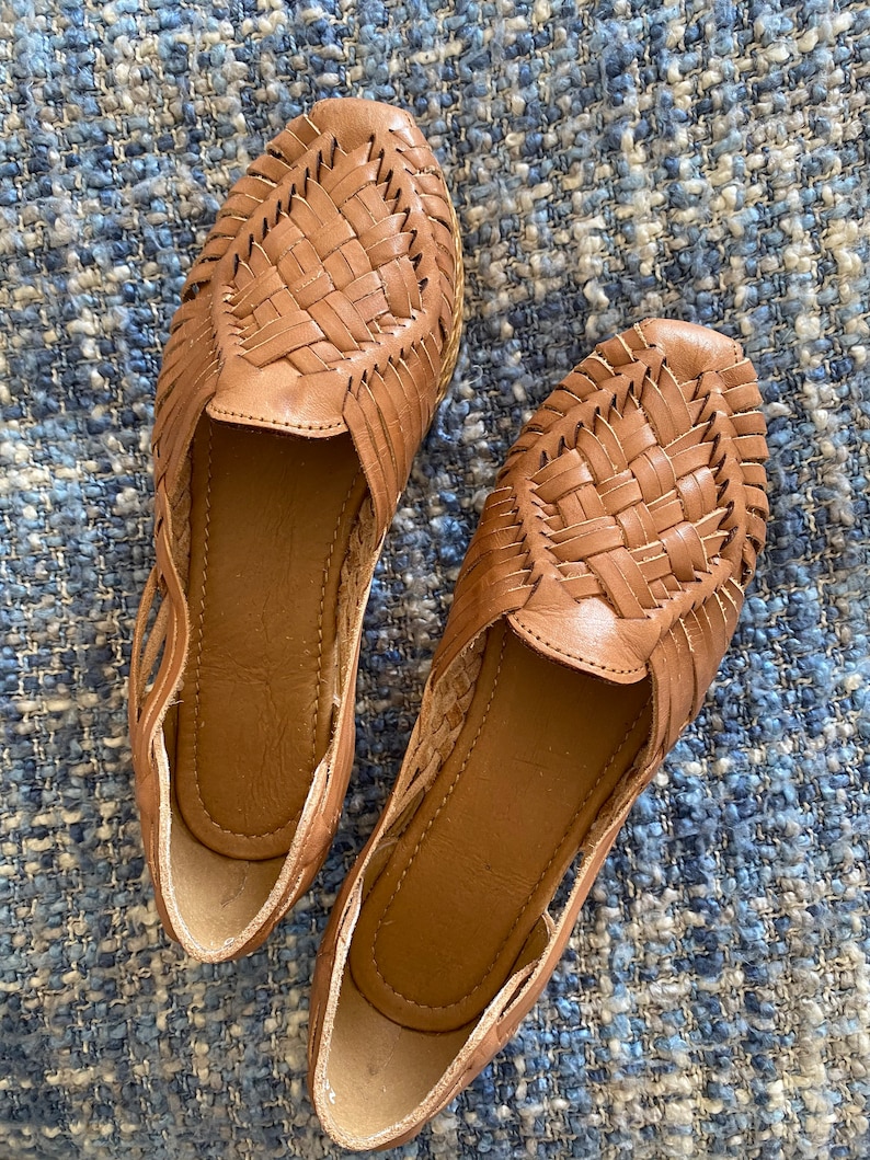 Op de afbeelding: Een paar bruine leren huarache-instappers met een gevlochten bovenwerk, een gesloten neus en een open zijkant. De schoenen hebben een gladde leren binnenzool en een flexibel, laag profiel met gedetailleerde handgeweven leren stroken.