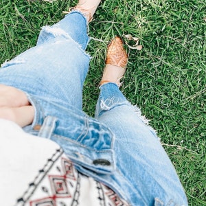 Puede incluir: Un par de sandalias huarache de cuero trenzado color canela con tiras finas en el tobillo, jeans de mezclilla azul claro desgastados con dobladillos deshilachados y una blusa blanca de manga larga con patr&oacute;n geom&eacute;trico de rombos negros y rojos.