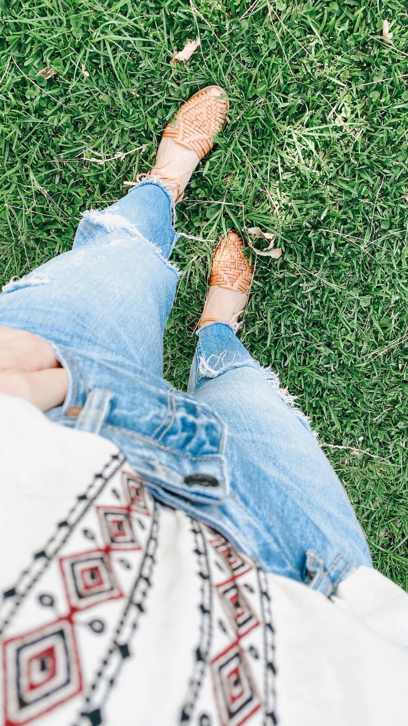 Puede incluir: Un par de sandalias huarache de cuero trenzado color canela con tiras finas en el tobillo, jeans de mezclilla azul claro desgastados con dobladillos deshilachados y una blusa blanca de manga larga con patr&oacute;n geom&eacute;trico de rombos negros y rojos.