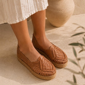 Op de afbeelding: Een paar instapschoenen in huarache-stijl van bruin leer met een gevlochten bovenwerk, een gesloten ronde neus en een dikke, getextureerde plateauzool van jute. De schoenen hebben een laag uitgesneden silhouet met uitsparingen aan de zijkanten.