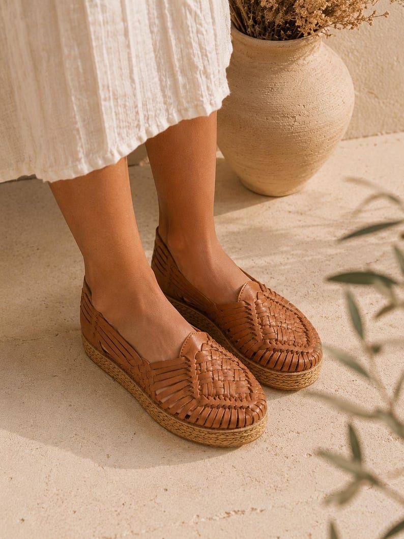 Op de afbeelding: Een paar instapschoenen in huarache-stijl van bruin leer met een gevlochten bovenwerk, een gesloten ronde neus en een dikke, getextureerde plateauzool van jute. De schoenen hebben een laag uitgesneden silhouet met uitsparingen aan de zijkanten.
