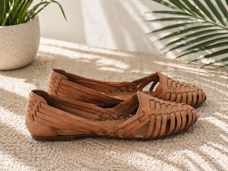 May include: A pair of tan huarache flats featuring a woven leather upper with a closed-toe design, an open-side construction, and a low heel. The shoes have a distinct interlaced pattern across the vamp and sides, providing a structured look.