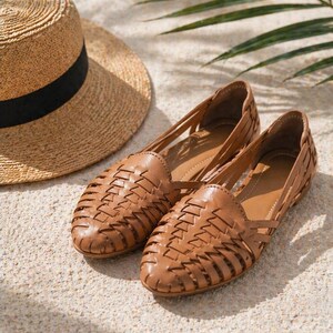 May include: A pair of tan leather huarache flats featuring a woven lattice design across the upper and a closed rounded toe, accompanied by a straw hat with a black ribbon band, showcasing a classic slip-on style with a low profile construction.