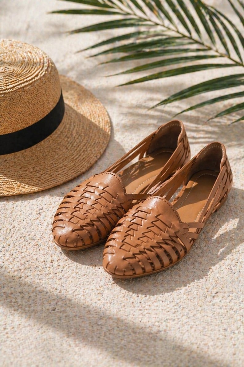 May include: A pair of tan leather huarache flats featuring a woven lattice design across the upper and a closed rounded toe, accompanied by a straw hat with a black ribbon band, showcasing a classic slip-on style with a low profile construction.