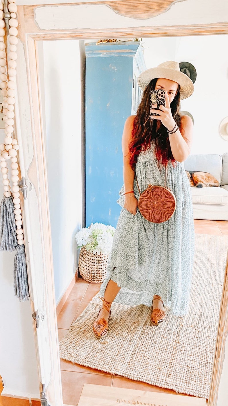 Puede incluir: Un vestido largo de color azul claro con estampado floral y silueta holgada, combinado con un sombrero de paja beige, un bolso bandolera redondo de rat&aacute;n trenzado con correa de cuero marr&oacute;n y sandalias huarache de cuero beige.