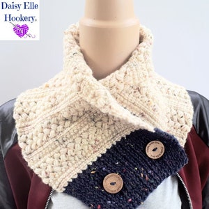 Puede incluir: Un cuello de punto de ganchillo en color crema y azul marino. El cuello tiene un patrón de punto texturizado y presenta dos botones de madera con el texto "Made with love" en ellos.