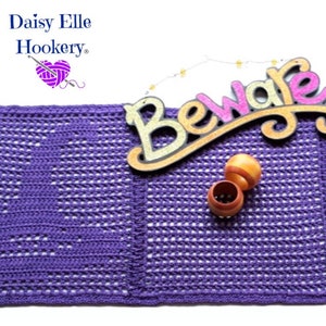 May include: Purple crochet table runner with three squares featuring a Halloween-themed design. The squares have a black cat, a pumpkin, and a skull design. The runner is decorated with a wooden sign that says "Beware".