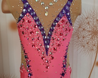 May include: Pink leotard with purple and clear rhinestones. The leotard has a V-neckline and a mesh overlay on the shoulders. The leotard is decorated with a pattern of rhinestones.
