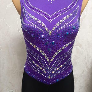 Puede incluir: Maillot morado sin mangas con cuello de malla y adornos de pedrería intrincados. El diseño presenta un patrón en forma de V de pedrería y una dispersión de gemas de colores más grandes. La parte inferior es negra.