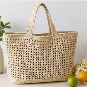 Könnte beinhalten: Eine beigefarbene Häkeltasche mit offenem Gewebe und zwei Griffen. Die Tasche ist rechteckig mit einem strukturierten Boden und einem großen, offenen Innenraum. Eine grüne Limette und eine Netztasche mit Zitronen liegen neben der Tasche.