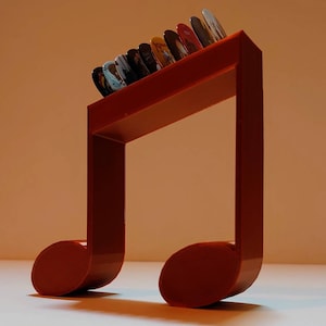 Puede incluir: Un soporte para p&uacute;as de guitarra de pl&aacute;stico rojo en forma de nota musical con una fila de p&uacute;as de colores en la parte superior.