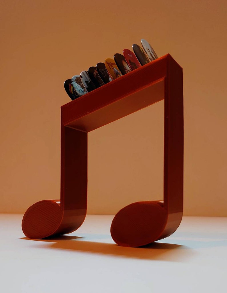 Puede incluir: Un soporte para p&uacute;as de guitarra de pl&aacute;stico rojo en forma de nota musical con una fila de p&uacute;as de colores en la parte superior.