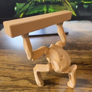 May include: A light tan, 3D-printed figurine depicting a stylized, mechanical-looking creature in a crouched position. The figure has two arms raised above its head, supporting a long, rectangular, solid block of the same material and color.