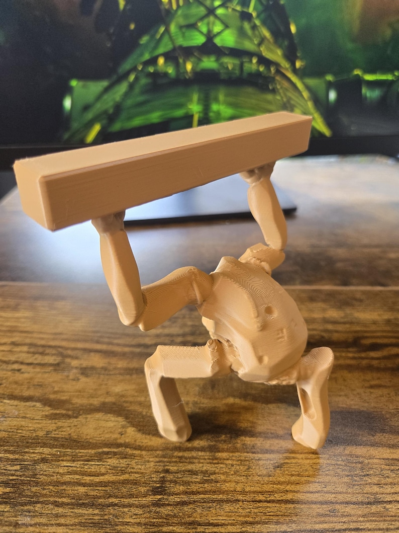 May include: A light tan, 3D-printed figurine depicting a stylized, mechanical-looking creature in a crouched position. The figure has two arms raised above its head, supporting a long, rectangular, solid block of the same material and color.