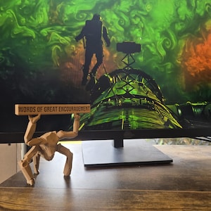 May include: A light brown, articulated, multi-legged creature figurine standing on a surface, holding a horizontal rectangular sign above its head with the black text "WORDS OF GREAT ENCOURAGEMENT" clearly printed on the front face of the sign.