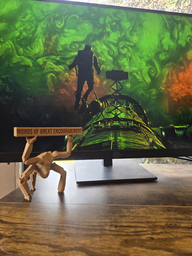 May include: A light brown, articulated, multi-legged creature figurine standing on a surface, holding a horizontal rectangular sign above its head with the black text "WORDS OF GREAT ENCOURAGEMENT" clearly printed on the front face of the sign.