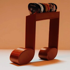 Puede incluir: Un soporte para p&uacute;as de guitarra de color marr&oacute;n impreso en 3D en forma de nota musical con una variedad de p&uacute;as. Las p&uacute;as tienen nombres de bandas y logotipos, incluyendo "Status Quo".