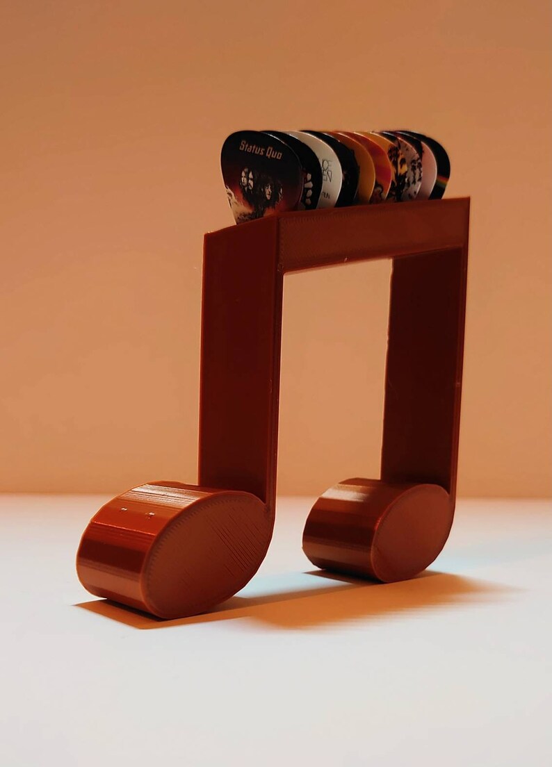Puede incluir: Un soporte para p&uacute;as de guitarra de color marr&oacute;n impreso en 3D en forma de nota musical con una variedad de p&uacute;as. Las p&uacute;as tienen nombres de bandas y logotipos, incluyendo "Status Quo".