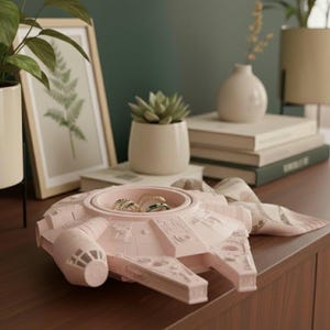 Könnte beinhalten: Ein hellrosa Schmuck-Organizer in Form des Raumschiffs Millennium Falcon. Der Organizer hat ein kreisförmiges Fach mit mehreren Ringen. Das Raumschiff steht auf einer dunkelbraunen Holzoberfläche.