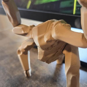 May include: A light tan, 3D-printed articulated figure with a humanoid torso and multiple limbs. The surface displays visible horizontal layer lines characteristic of additive manufacturing, with jointed segments allowing for various poses.