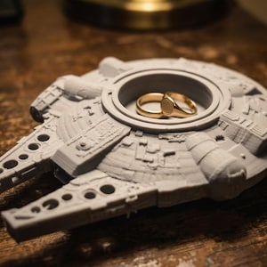 Op de afbeelding: Een grijze, 3D-geprinte Millennium Falcon-ringhouder met twee gouden ringen erin. De ringhouder is gedetailleerd met verschillende vormen en texturen en ligt op een houten oppervlak.