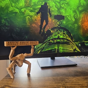 May include: A light tan, articulated, multi-legged figure with a blocky, insect-like appearance, holding a rectangular sign above its head that features the repeated text "AMAZE AMAZE AMAZE" in dark, embossed lettering on its front surface.