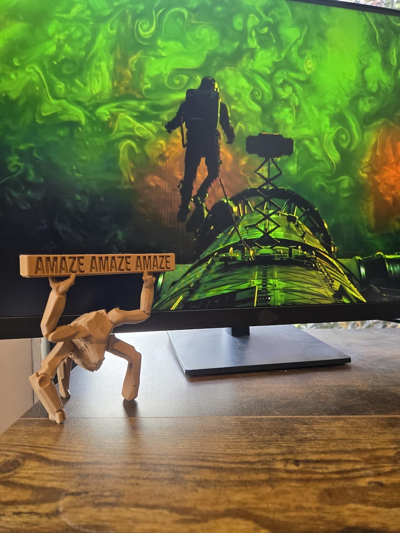 May include: A light tan, articulated, multi-legged figure with a blocky, insect-like appearance, holding a rectangular sign above its head that features the repeated text "AMAZE AMAZE AMAZE" in dark, embossed lettering on its front surface.