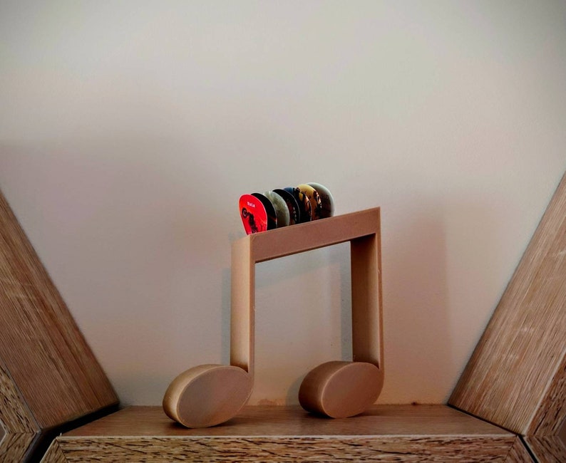 Puede incluir: Un soporte de madera para p&uacute;as de guitarra con forma de nota musical. El soporte es marr&oacute;n y tiene algunas p&uacute;as de guitarra en &eacute;l.