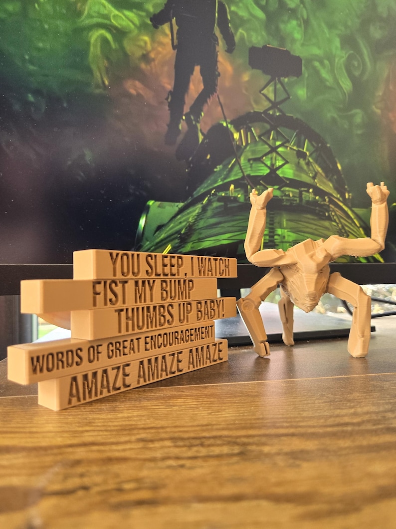 May include: A beige, multi-tiered block structure featuring engraved text that reads: YOU SLEEP, I WATCH, FIST MY BUMP, THUMBS UP BABY!, WORDS OF GREAT ENCOURAGEMENT, and AMAZE AMAZE AMAZE. Beside it stands a beige, articulated, multi-legged creature figurine.