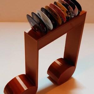 Puede incluir: Un soporte para p&uacute;as de guitarra de pl&aacute;stico marr&oacute;n en forma de nota musical con una fila de p&uacute;as de colores en la parte superior.